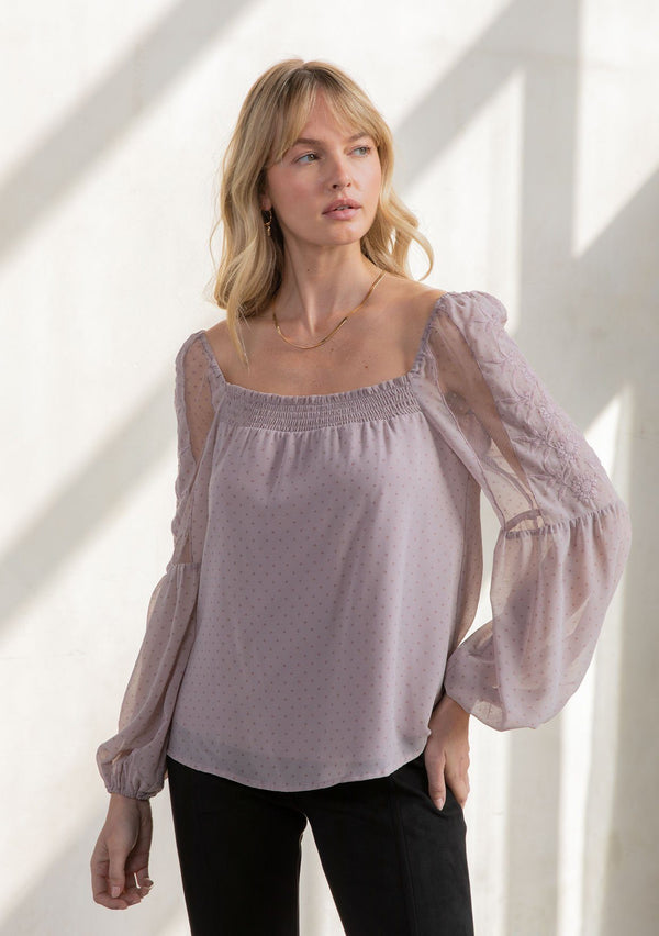 [Color: Vintage Lilac] A model wearing a romantic square neck blouse in a lilac purple dot chiffon. With a long voluminous lantern sleeve with embroidered dot mesh detail and a billowy relaxed fit.