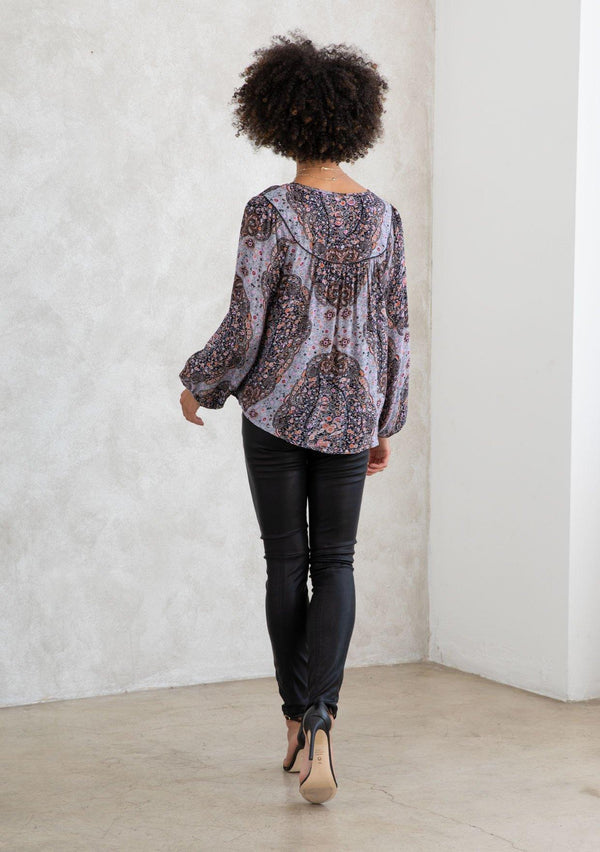 [Color: Grey/Rust] A model wearing a grey and rust paisley print bohemian peasant top. A classic style with long voluminous sleeves, a split v neckline with tassel ties, and a bib yoke detail. 