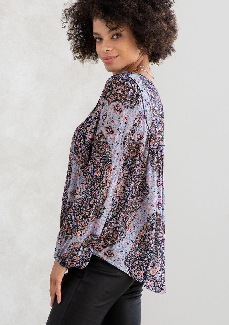 [Color: Grey/Rust] A model wearing a grey and rust paisley print bohemian peasant top. A classic style with long voluminous sleeves, a split v neckline with tassel ties, and a bib yoke detail. 