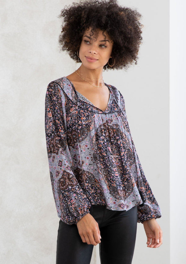 [Color: Grey/Rust] A model wearing a grey and rust paisley print bohemian peasant top. A classic style with long voluminous sleeves, a split v neckline with tassel ties, and a bib yoke detail. 