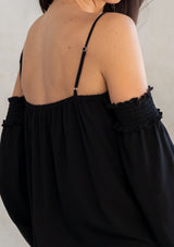 [Color: Black] A model wearing an ultra bohemian billowy black blouse with long attached sleeves, an elastic neckline, and adjustable spaghetti straps. 