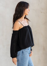 [Color: Black] A model wearing an ultra bohemian billowy black blouse with long attached sleeves, an elastic neckline, and adjustable spaghetti straps. 