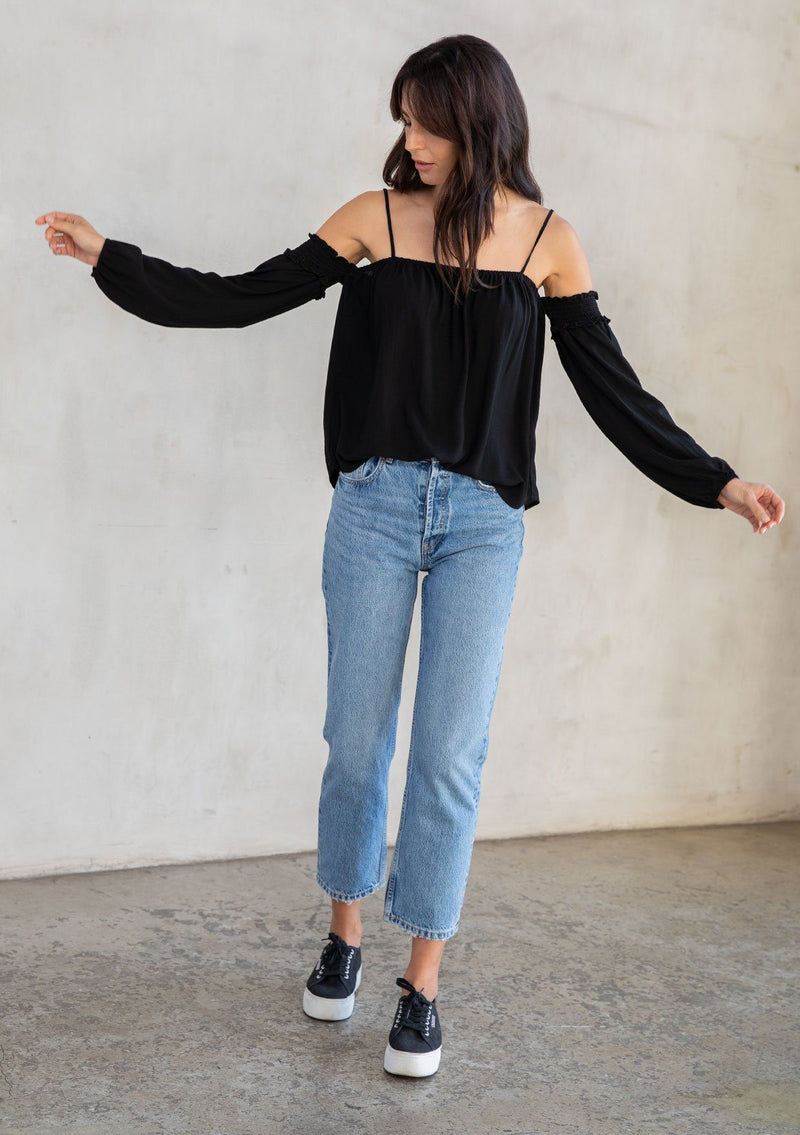 [Color: Black] A model wearing an ultra bohemian billowy black blouse with long attached sleeves, an elastic neckline, and adjustable spaghetti straps. 