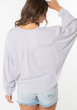 [Color: Dusty Lilac] A model wearing a flowy long sleeve light purple bohemian tie front top.