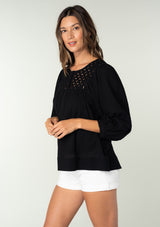 [Color: Black] A side facing image of a brunette model wearing a black cotton bohemian blouse. With long puff sleeves, a flowy relaxed fit, and a lattice front yoke.