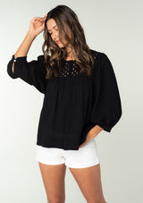 [Color: Black] A front facing image of a brunette model wearing a black cotton bohemian blouse. With long puff sleeves, a flowy relaxed fit, and a lattice front yoke.