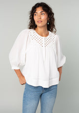 [Color: White] A front facing image of a brunette model wearing a white cotton bohemian blouse. With long puff sleeves, a flowy relaxed fit, and a lattice front yoke. 