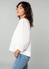 [Color: White] A side facing image of a brunette model wearing a white cotton bohemian blouse. With long puff sleeves, a flowy relaxed fit, and a lattice front yoke. 