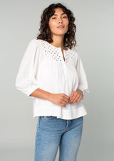 [Color: White] A half body front facing image of a brunette model wearing a white cotton bohemian blouse. With long puff sleeves, a flowy relaxed fit, and a lattice front yoke. 