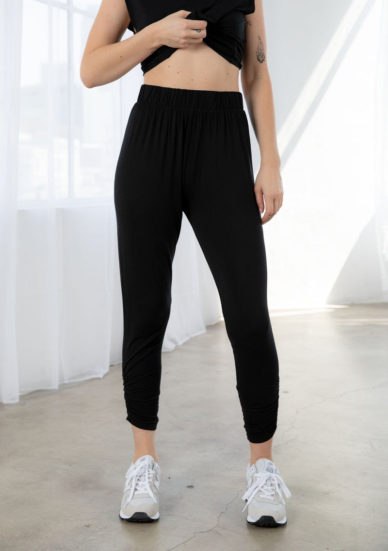 [Color: Black] A model wearing a soft and stretchy legging pant. With a cropped leg, side gathered details at the ankle, and an elastic waist.