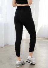 [Color: Black] A model wearing a soft and stretchy legging pant. With a cropped leg, side gathered details at the ankle, and an elastic waist.