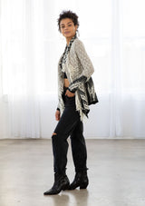 [Color: Light Heather Grey/Black] A model wearing a chunky marled knit bohemian cardigan in a grey and black pattern. With a fringed hemline, long sleeves, and contrast trim.