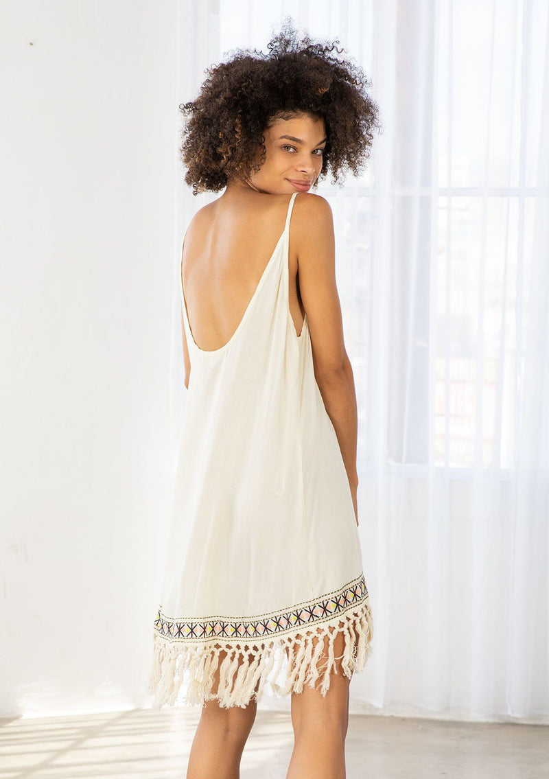 [Color: Natural] A model wearing a natural off white sleeveless mini dress with a scooped neckline, low scooped back, and fringed hemline.