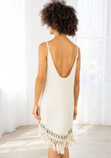 [Color: Natural] A model wearing a natural off white sleeveless mini dress with a scooped neckline, low scooped back, and fringed hemline.