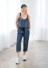 [Color: Midnight Wash] A blond model wearing a denim blue classic jumpsuit made from eco friendly Tencel. Featuring a wide leg with a cuffed hem, essential side pockets, and a button front.