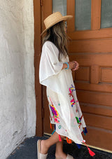 [Color: Off White] A brunette woman standing outside wearing a mid length sheer caftan with bold embroidered detail and colorful tassel trim.
