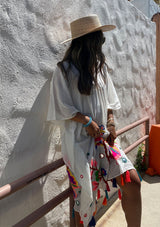 [Color: Off White] A brunette woman standing outside wearing a mid length sheer caftan with bold embroidered detail and colorful tassel trim.