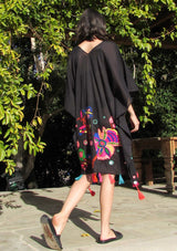 [Color: Black] A brunette woman standing poolside wearing a mid length sheer caftan with bold embroidered detail and colorful tassel trim.