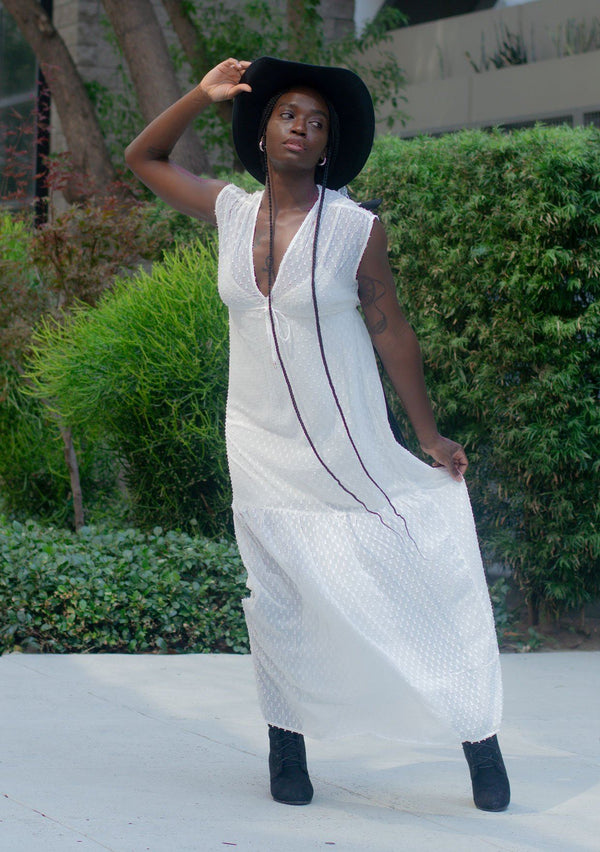 [Color: Off White] A woman wearing a sheer off white chiffon swiss dot maxi dress. With a drawstring empire waist, tiered skirt, v neckline, and lining. 