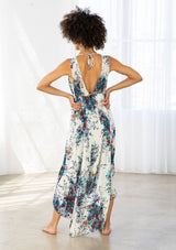 [Color: Natural/Turquoise] A model wearing a natural and blue watercolor floral print maxi dress. With a high low hemline, a tiered skirt, a v neckline, an empire waist with front tie, and an open back with tie closure. 