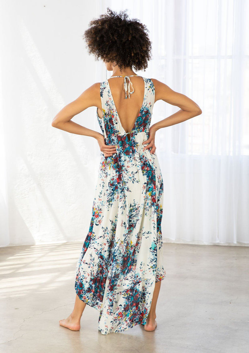 [Color: Natural/Turquoise] A model wearing a natural and blue watercolor floral print maxi dress. With a high low hemline, a tiered skirt, a v neckline, an empire waist with front tie, and an open back with tie closure. 