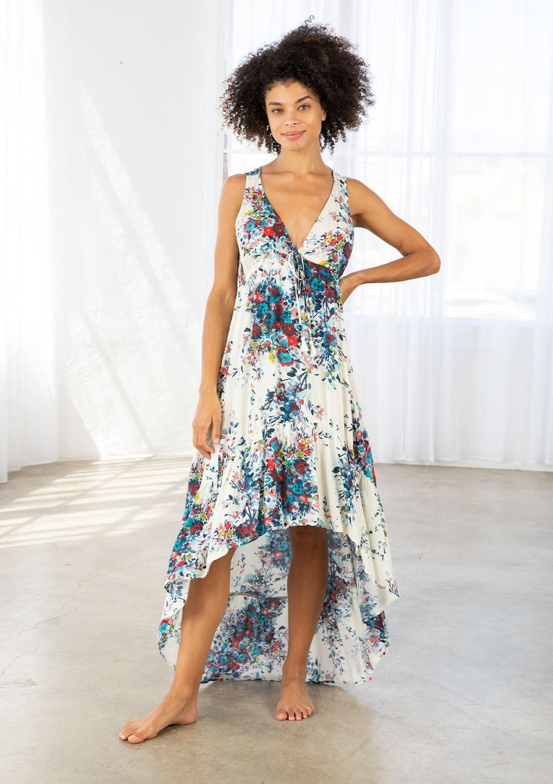 [Color: Natural/Turquoise] A model wearing a natural and blue watercolor floral print maxi dress. With a high low hemline, a tiered skirt, a v neckline, an empire waist with front tie, and an open back with tie closure. 