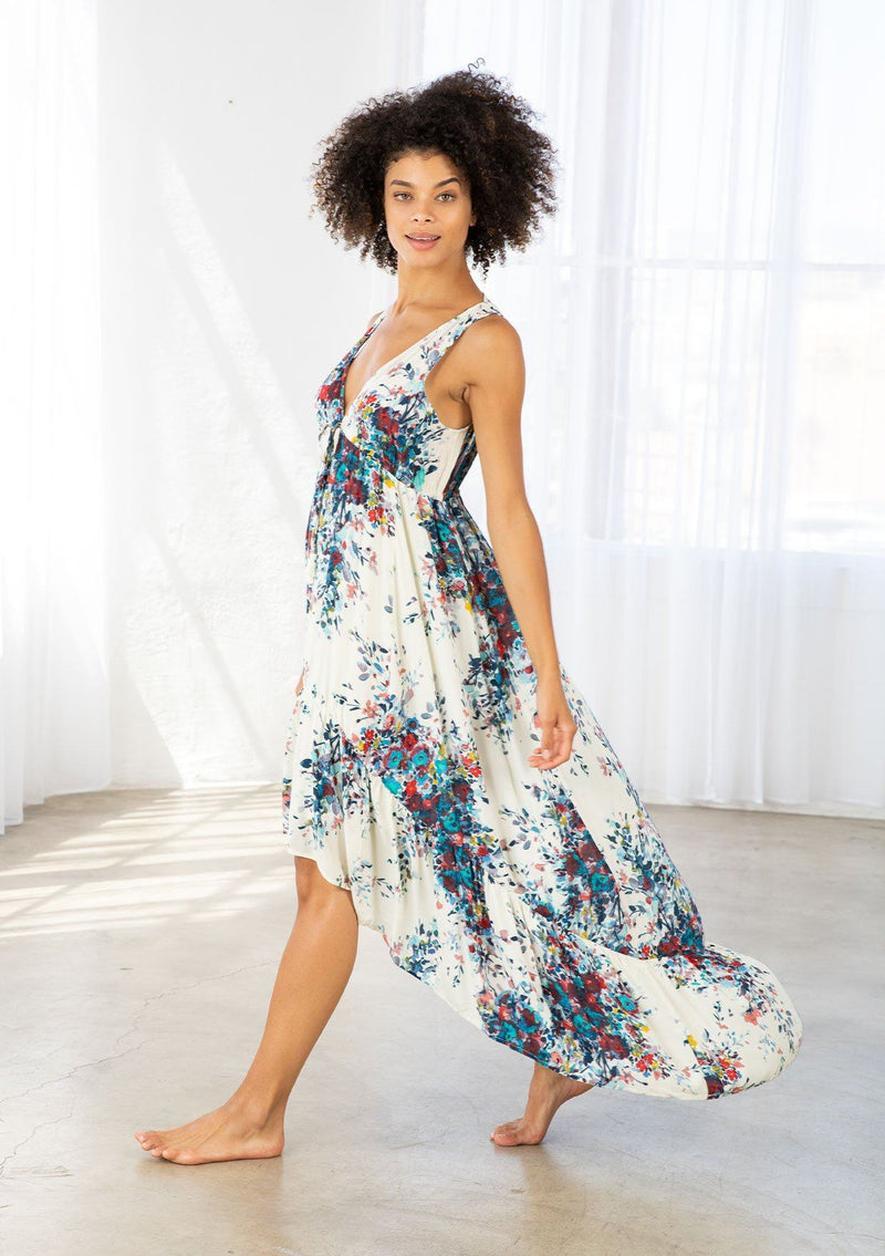 [Color: Natural/Turquoise] A model wearing a natural and blue watercolor floral print maxi dress. With a high low hemline, a tiered skirt, a v neckline, an empire waist with front tie, and an open back with tie closure. 