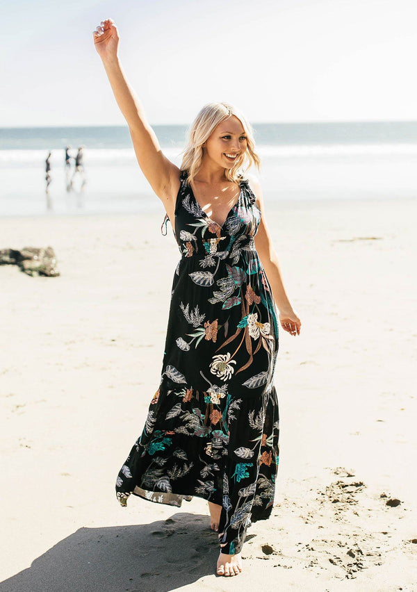 [Color: Black Combo] A woman standing on the beach wearing a large floral print sleeveless maxi dress. With an empire waist, a v neckline in front and back, and tie shoulder details. 