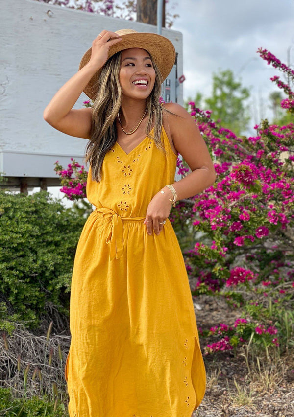 [Color: Mustard] A woman standing outside wearing a bright mustard yellow sleeveless mid length dress. Featuring a v neckline, a self tie waist, side slits, and delicate eyelet details. 