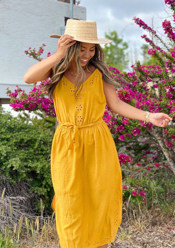 [Color: Mustard] A woman standing outside wearing a bright mustard yellow sleeveless mid length dress. Featuring a v neckline, a self tie waist, side slits, and delicate eyelet details. 