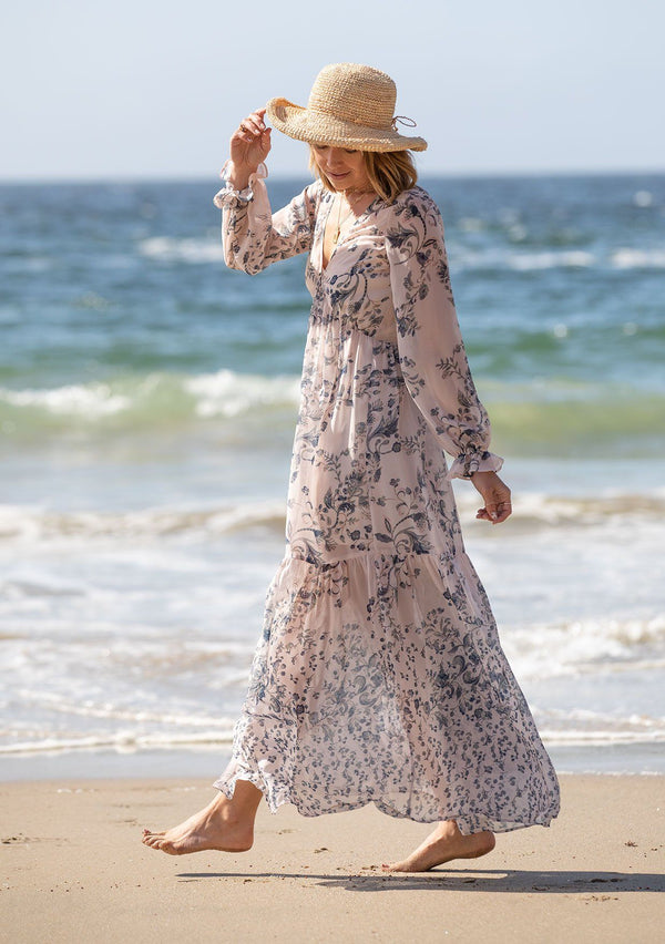 [Color: Light Peach] A woman on the beach wearing a dreamy vintage floral maxi dress. Featuring long voluminous sheer sleeves with a flounce wrist cuff, a button front, and an empire waist.