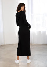 [Color: Black] A model wearing a slim fit black maxi dress in stretchy rib. With long sleeves, thumb holes, a hoodie, and a side slit. 