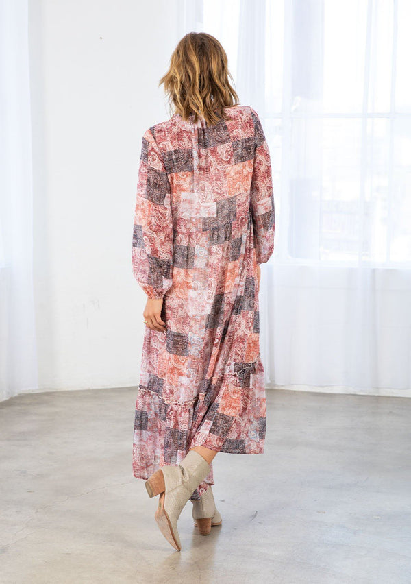 [Color: Natural/Rose] A model wearing a loose and billowy bohemian maxi dress in a red paisley patchwork print. With long voluminous sleeves, a button front, a ruffle trimmed neckline, and a tiered skirt.