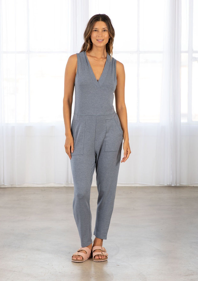 [Color: Denim Blue] A model wearing a blue soft French terry sleeveless jumpsuit. With a v neckline, side patch pockets, a long tapered leg, and contrast ribbed trim detail. 