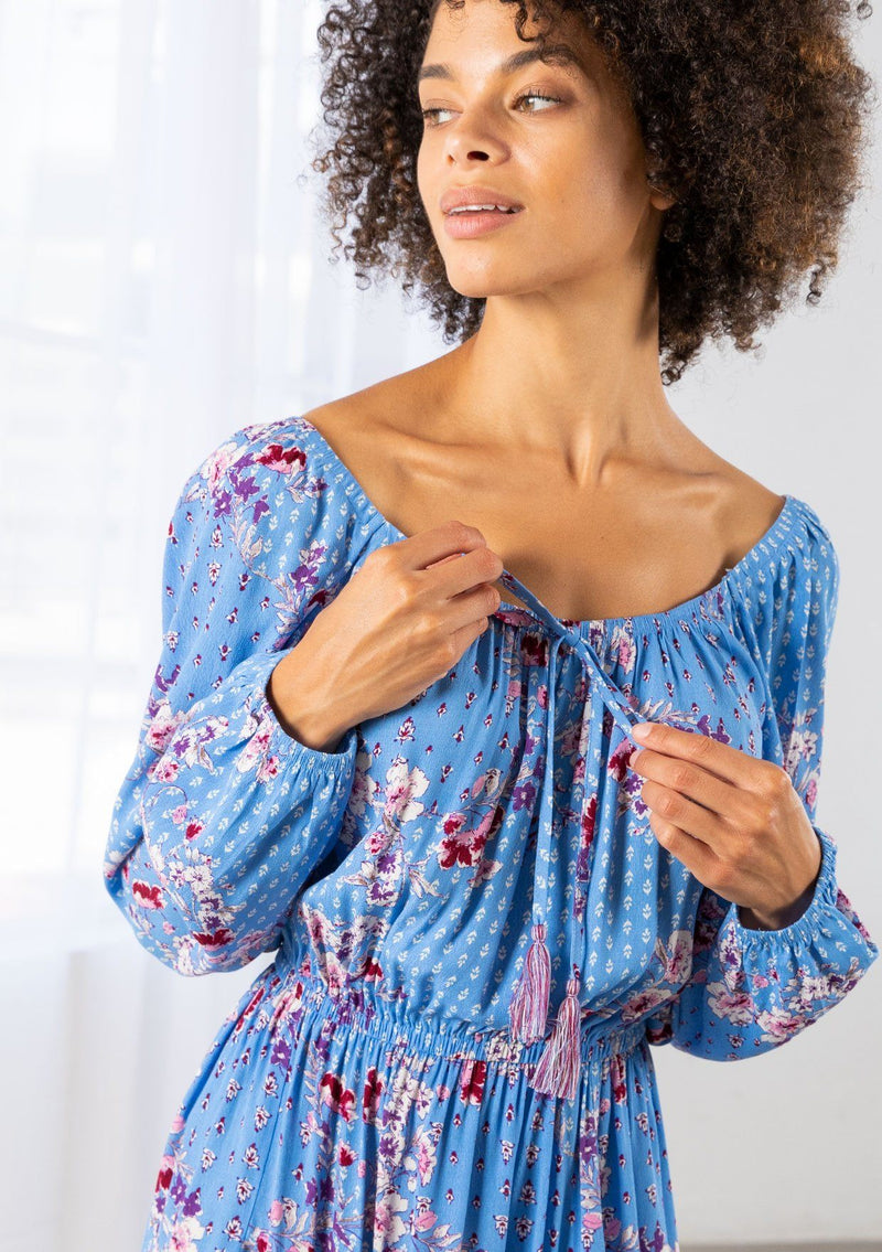 [Color: Azure/Mulberry] A model wearing a timeless bohemian mini dress in a vibrant blue floral print. With long voluminous sleeves and an elastic wrist cuff, a tiered skirt, a front tassel tie, an elastic waist, and a round elastic neckline that can be worn on or off the shoulder. 