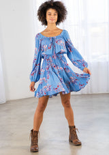 [Color: Azure/Mulberry] A model wearing a timeless bohemian mini dress in a vibrant blue floral print. With long voluminous sleeves and an elastic wrist cuff, a tiered skirt, a front tassel tie, an elastic waist, and a round elastic neckline that can be worn on or off the shoulder. 