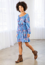 [Color: Azure/Mulberry] A model wearing a timeless bohemian mini dress in a vibrant blue floral print. With long voluminous sleeves and an elastic wrist cuff, a tiered skirt, a front tassel tie, an elastic waist, and a round elastic neckline that can be worn on or off the shoulder. 