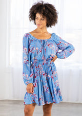 [Color: Azure/Mulberry] A model wearing a timeless bohemian mini dress in a vibrant blue floral print. With long voluminous sleeves and an elastic wrist cuff, a tiered skirt, a front tassel tie, an elastic waist, and a round elastic neckline that can be worn on or off the shoulder.  