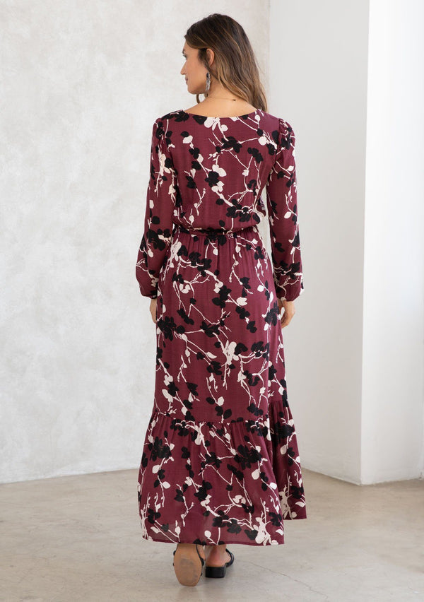 [Color: Merlot/Ivory] A model wearing a merlot red blossom floral print maxi dress. With a mini pom trimmed v neckline, an elastic waist, a side slit, voluminous long sleeves with an elastic wrist cuff, and attached lining. 