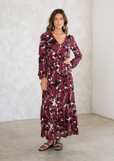 [Color: Merlot/Ivory] A model wearing a merlot red blossom floral print maxi dress. With a mini pom trimmed v neckline, an elastic waist, a side slit, voluminous long sleeves with an elastic wrist cuff, and attached lining. 