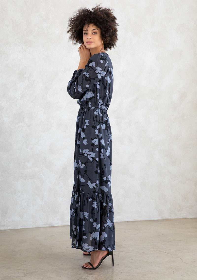 [Color: Slate/Grey] A model wearing a slate blue and grey blossom floral print maxi dress. With a mini pom trimmed v neckline, an elastic waist, a side slit, voluminous long sleeves with an elastic wrist cuff, and attached lining. 