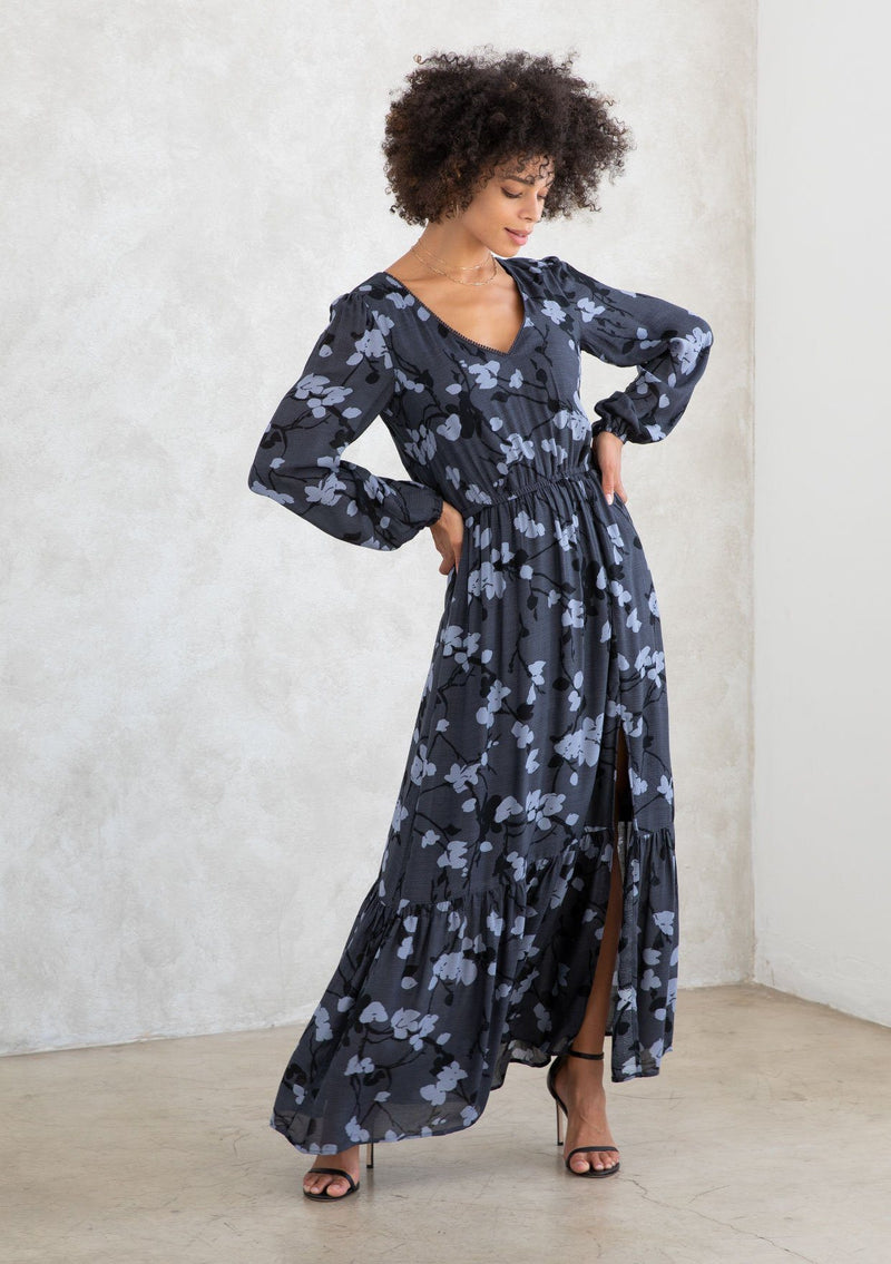 [Color: Slate/Grey] A model wearing a slate blue and grey blossom floral print maxi dress. With a mini pom trimmed v neckline, an elastic waist, a side slit, voluminous long sleeves with an elastic wrist cuff, and attached lining. 