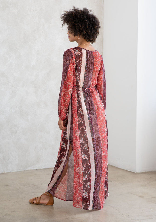 [Color: Brick Red/Plum] A model wearing a sheer red and purple maxi dress in a bohemian floral patchwork print. With long voluminous sleeves, a loop button front, and side slits. 