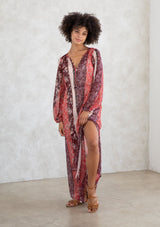 [Color: Brick Red/Plum] A model wearing a sheer red and purple maxi dress in a bohemian floral patchwork print. With long voluminous sleeves, a loop button front, and side slits. 