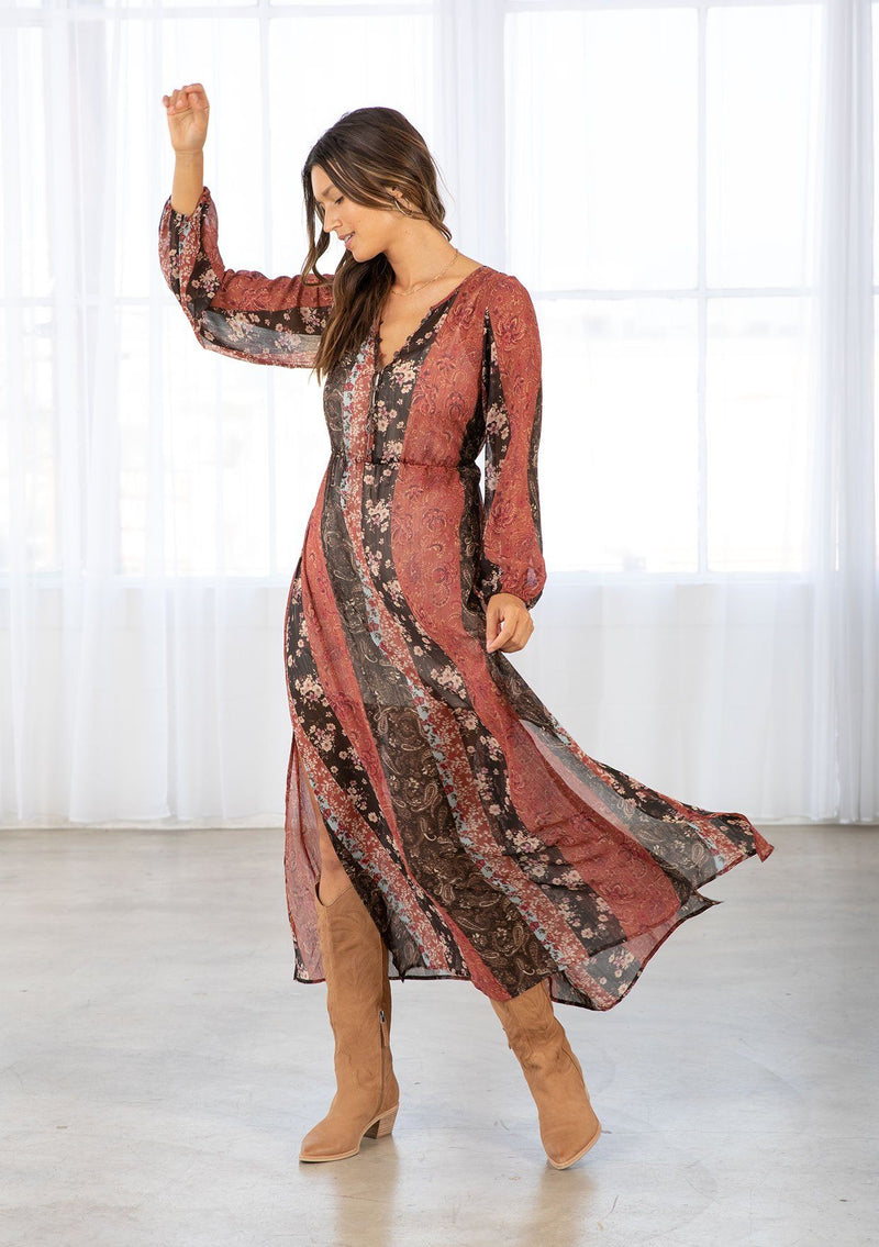 [Color: Cinnamon/Brown] A model wearing a sheer red and brown maxi dress in a bohemian floral patchwork print. With long voluminous sleeves, a loop button front, and side slits. 