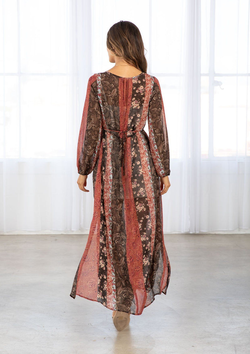 [Color: Cinnamon/Brown] A model wearing a sheer red and brown maxi dress in a bohemian floral patchwork print. With long voluminous sleeves, a loop button front, and side slits. 