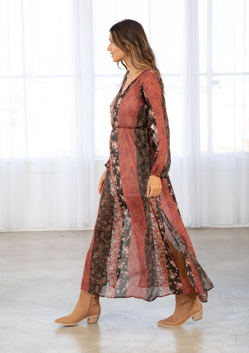 [Color: Cinnamon/Brown] A model wearing a sheer red and brown maxi dress in a bohemian floral patchwork print. With long voluminous sleeves, a loop button front, and side slits. 