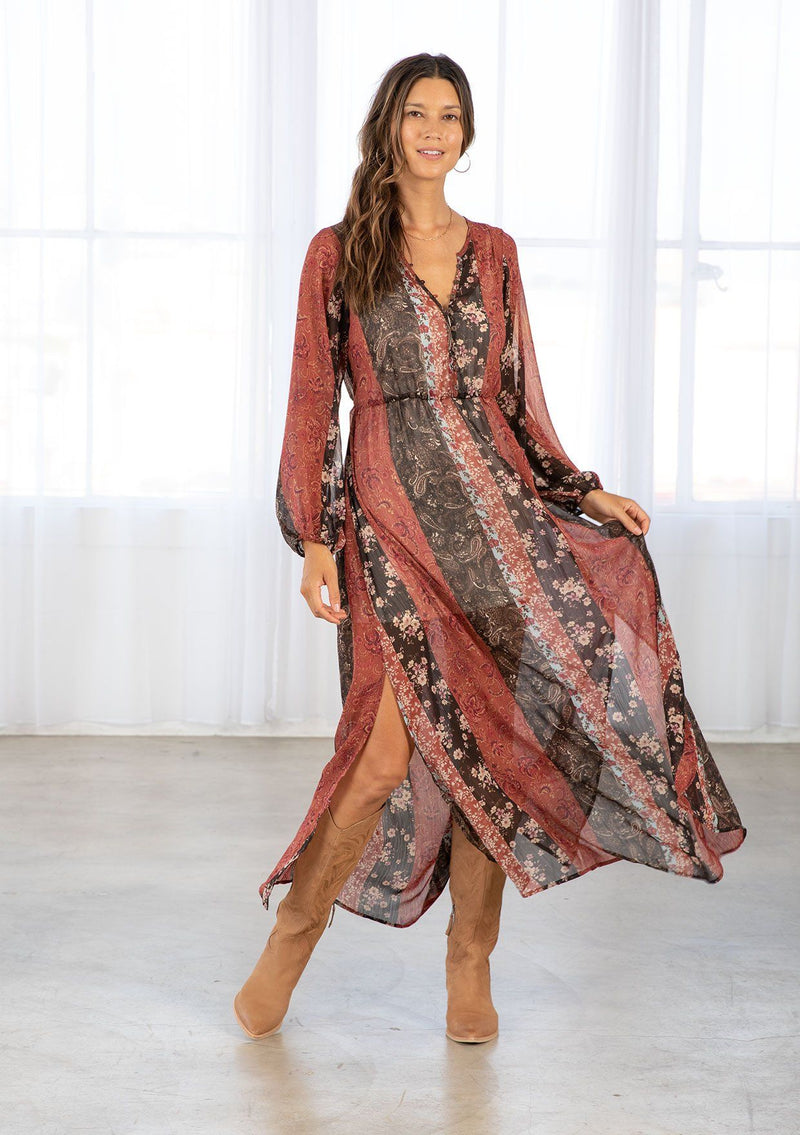 [Color: Cinnamon/Brown] A model wearing a sheer red and brown maxi dress in a bohemian floral patchwork print. With long voluminous sleeves, a loop button front, and side slits. 