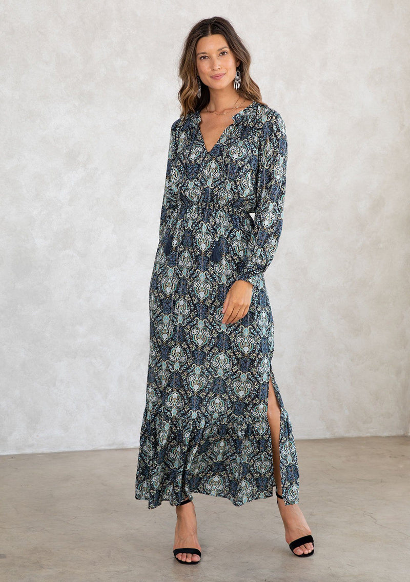 [Color: Black/Teal] A model wearing a black and teal paisley print maxi dress with light catching lurex thread. With long voluminous sleeves, a split neckline with tassel ties, side slits, and an elastic waist. 