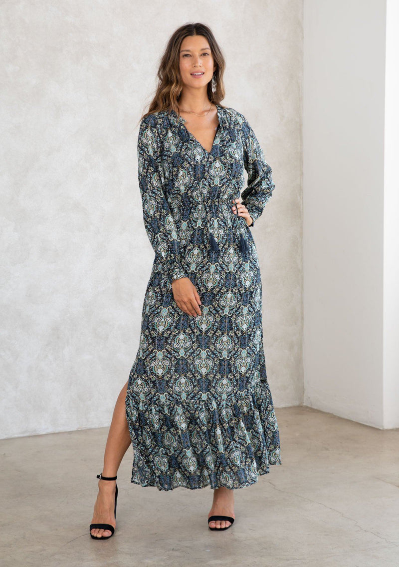 [Color: Black/Teal] A model wearing a black and teal paisley print maxi dress with light catching lurex thread. With long voluminous sleeves, a split neckline with tassel ties, side slits, and an elastic waist. 